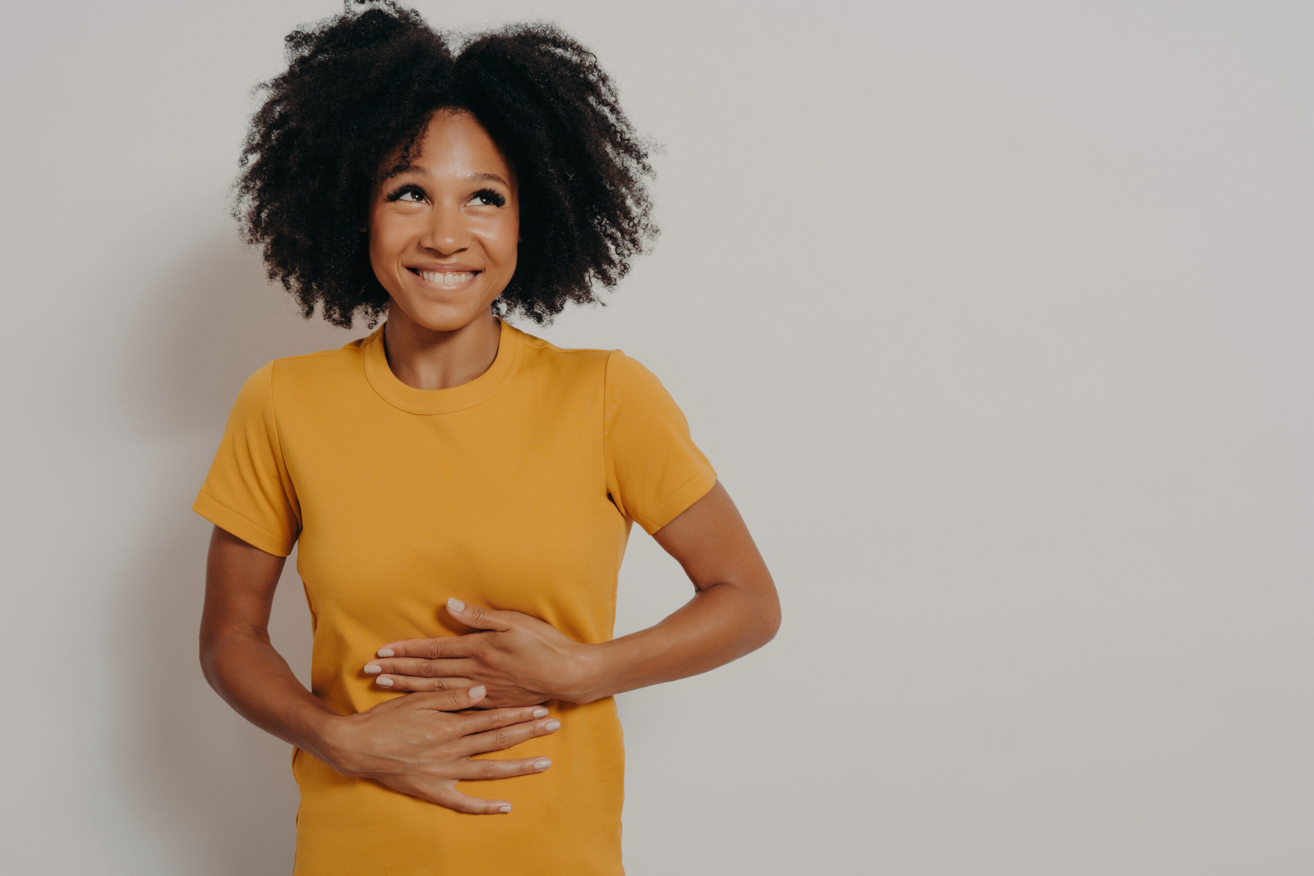 A woman with curly hair, wearing a yellow t-shirt, smiles and looks up while placing both hands on her stomach against a plain white background.