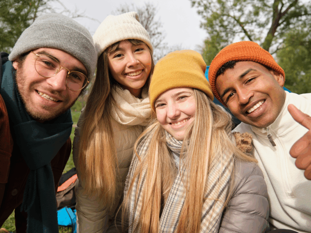 Four friends discussing the gut and immune system, wearing winter hats and jackets smile and pose closely together outdoors, surrounded by trees and greenery on a chilly day. One person gives a thumbs up.
