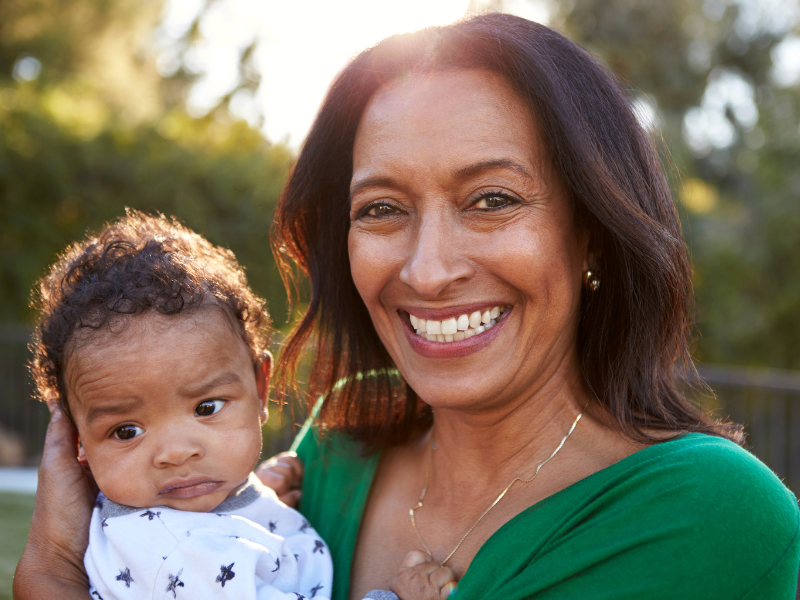 A smiling woman in a green top holds a baby wearing a star-patterned onesie outdoors. The sun shines behind them, and greenery is visible in the background.